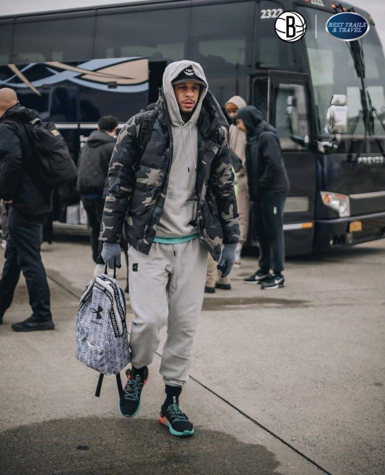 Person Wearing A Camouflage Puffer Jacket, A Light Grey Hoodie, Light Grey Sweatpants, And Black And Green Athletic Shoes Walks Across An Asphalt Lot, Carrying A Grey And White Patterned Backpack In One Hand